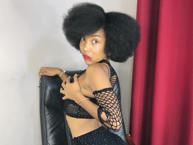 TynnaMagik with big afro, red lipstick, wearing black fishnet top and bottom, covering small tits with left hand, standing against black leather chair, red curtain on right.