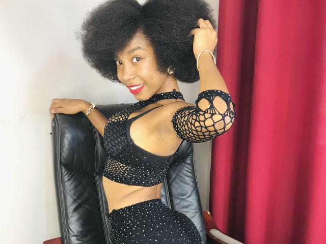 TynnaMagik with big afro, smiling, wearing black mesh crop top and skirt with sparkles, holding chair, red curtains, black leather chair, red lipstick, gold bracelet.
