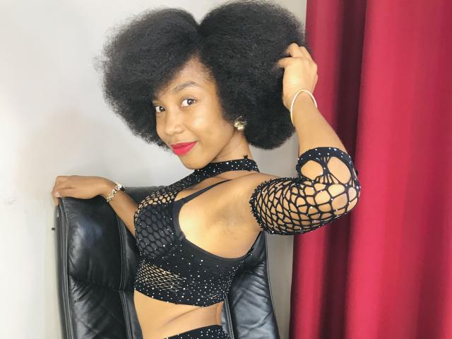 TynnaMagik with big afro, red lipstick, black fishnet top, exposing small tits, holding hair, sitting on black leather chair, red curtain on right, smiling at camera.