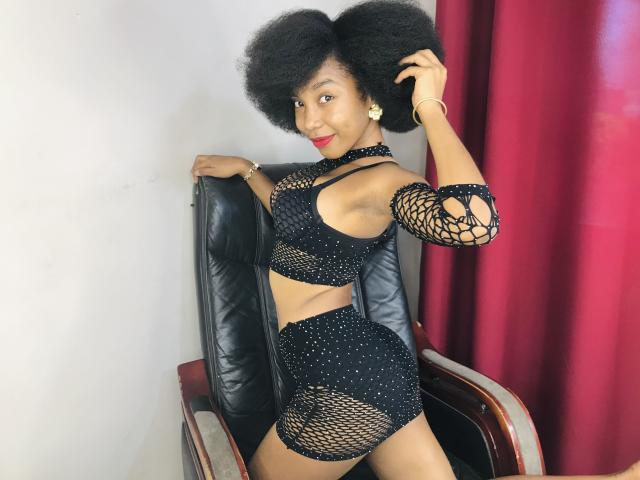TynnaMagik with a big afro, wearing a black fishnet outfit that exposes her tits and ass, standing in a chair, smiling, touching her hair, red curtains in background.