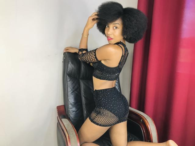 TynnaMagik with big black afro, wearing a black mesh dress showing her ass and tits, kneeling on a leather chair, red curtains in the background, looking back.