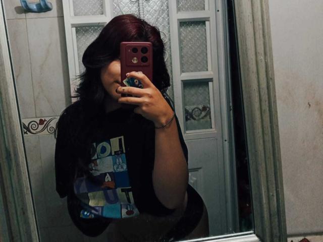 Milla Casanova met rood haar, gekleed in een zwart shirt met kleurrijke vierkantjes, maakt een spiegelzelfportret met een rode telefoon, waarbij haar zwart gelakte nagels en een armband te zien zijn..