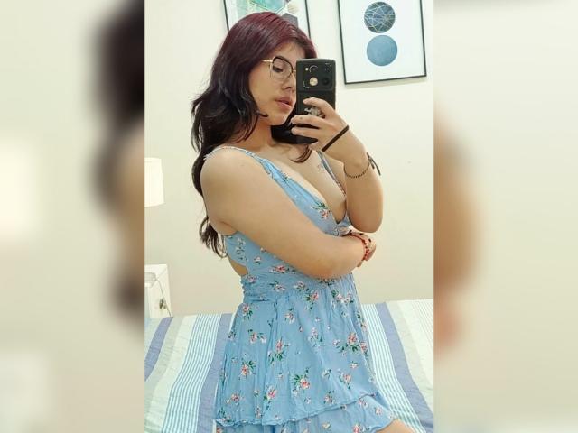 MillaCasanova with red hair, glasses, and a blue floral dress, showing her tits in a mirror selfie, holding a phone, in a bright room.