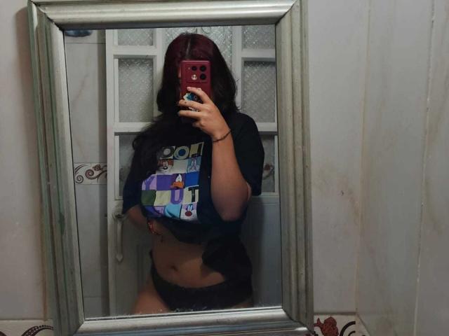 Selfie en el espejo de MillaCasanova con cabello oscuro, vistiendo una camiseta negra y ropa interior negra, mostrando su abdomen descubierto y sus pequeños senos, sosteniendo un teléfono rojo..