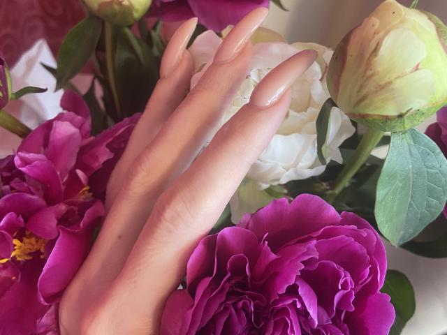Nalini's hand with long, nude-painted nails rests on vibrant pink and white peonies, showing off her slender fingers and fair skin. Her ass is not visible.