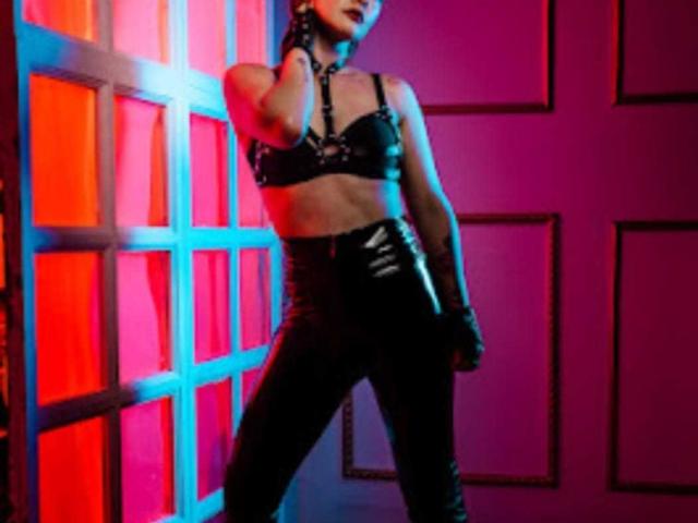 Antomouth stands in black leather lingerie with shiny pants, a choker, and gloves, illuminated by pink and blue lights, showing her slim waist and small tits.