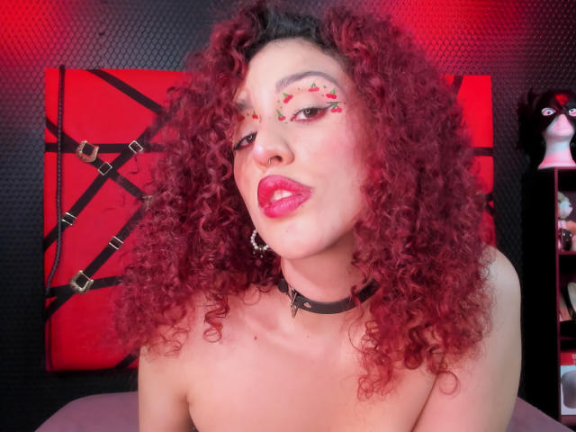 KaylinKrof with curly red hair, red lipstick, and face paint, wearing a black choker, looking at the camera with a pouty expression, showing her bare shoulders.
