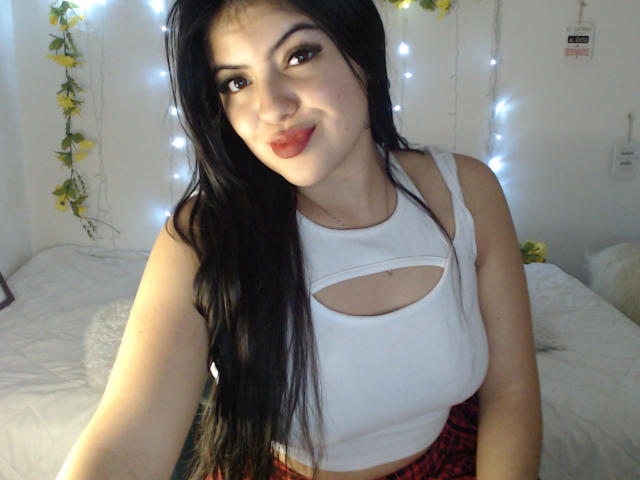 AlissonS has long black hair, wearing a white cutout top, showing small tits, red plaid skirt, and red lipstick, smiling in a bedroom with string lights.