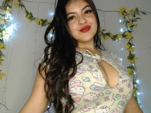 AlissonS with long black hair, red lipstick, and large tits in a Hello Kitty-patterned top, smiling at the camera. Yellow flowers and white string lights in the background.