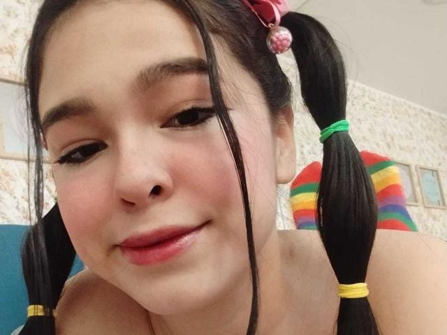 DulceTernura met vlechtjes, een roze strik, en een achtergrond met regenboogstrepen, lachend met roze lippen, zwarte eyeliner, en gele haarbandjes, waarbij haar schattige gezicht en subtiele make-up te zien zijn..