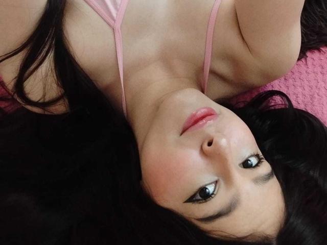 Le visage de DulceTernura, allongée, avec ses cheveux noirs, ses bretelles de soutien-gorge roses et un fond texturé rose, montrant ses lèvres charnues et ses yeux marrons, regardant vers l'appareil photo..