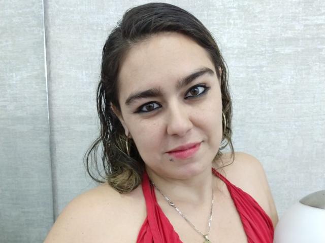 TaylorNova mit feuchtem, braunem Haar, einem roten Top mit Halter, schwarzem Eyeliner, goldenen Creolen, einem kleinen Nasenpiercing, einer silbernen Halskette und einem leichten Lächeln, vor einer grauen, strukturierten Wand..