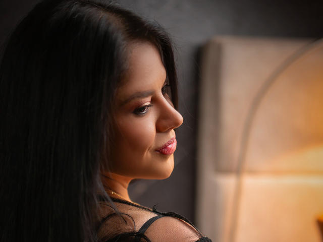A sexy photo of MilfMonserrat with long black hair, smiling at the camera, showing her cute nose and pink lips, wearing a black sheer top, and a gold necklace.