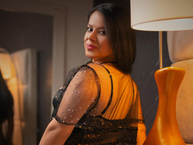 MilfMonserrat with dark hair, smiling, wearing a black sheer top with polka dots, showing her back and looking over her shoulder, standing next to an orange lamp.