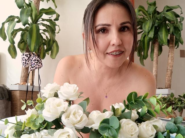AdelaShery, con ojos marrones, sonriendo, sosteniendo rosas blancas, sin ropa en la parte superior, usando un collar púrpura, plantas en macetas al fondo, mirando a la cámara, cabello castaño en una coleta..