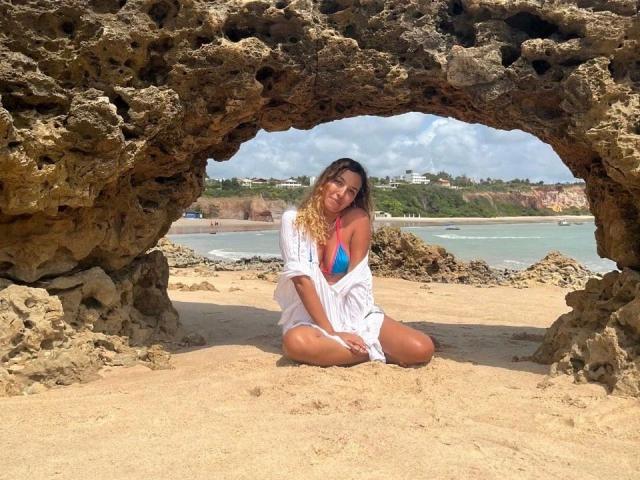 Sukievee, avec ses cheveux blonds bouclés, assise sur une plage de sable, portant une chemise blanche ouverte, un haut de bikini bleu et rose, montrant ses seins, encadrée par une grotte rocheuse, a l'air adorable, c'est une journée ensoleillée..