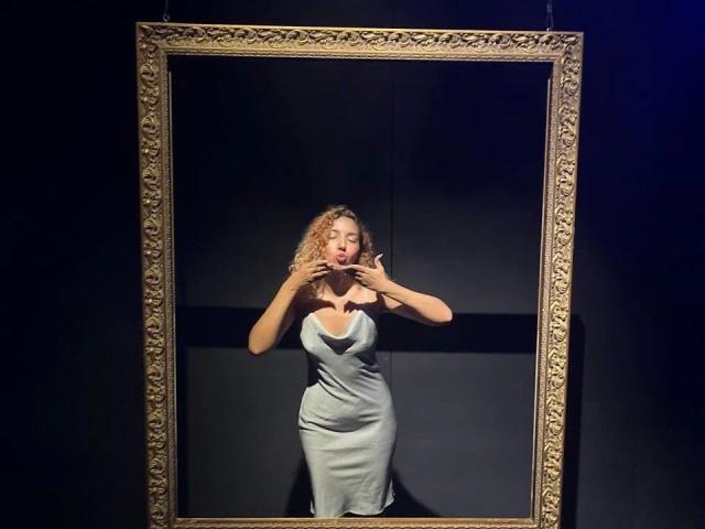 Sukievee-hot está de pie frente a un elaborado marco dorado, vestida con un vestido azul claro ajustado, con las manos cerca de sus pechos, con cabello rubio rizado y una sonrisa segura y sensual..