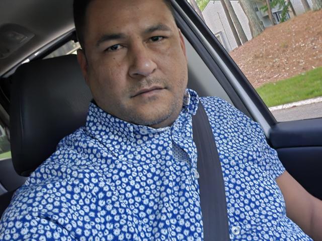 Yeiold, een dikkerd met kort zwart haar, gekleed in een blauw shirt met witte patronen, zit in een auto met een veiligheidsgordel en kijkt er enigszins verveeld uitzien..