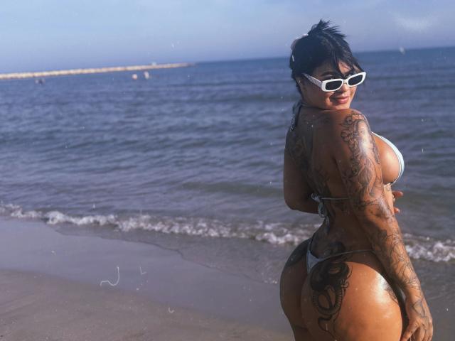Cloerhet, abbronzata e con tatuaggi, in bikini bianco, con occhiali da sole bianchi, capelli neri, pelle abbronzata, grande sedere, seno prosperoso, in piedi sulla spiaggia, che si guarda indietro, sorridendo, con l'oceano sullo sfondo..