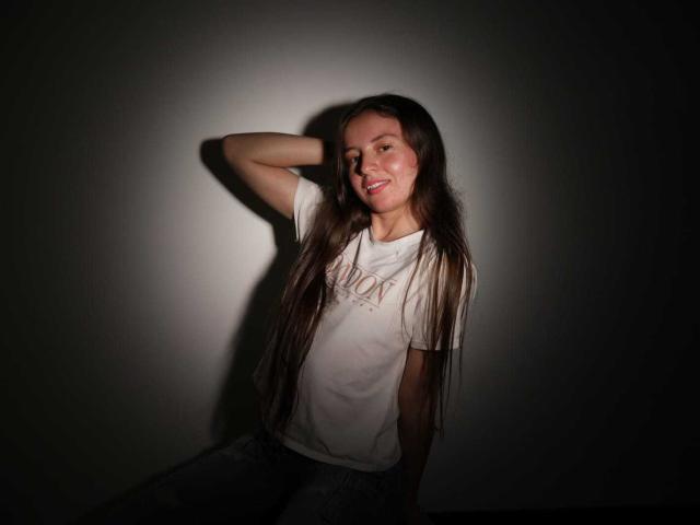 VioletaNova with long brown hair, white t-shirt, and blue jeans, smiling, arm behind head, shadowed against wall. Her tits are small, and she's sexy.