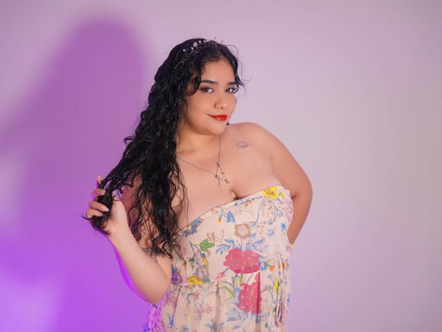 AliciaRelas with curly black hair, wearing a floral strapless dress, holding hair, red lipstick, tattoo on right shoulder, purple and pink lighting, slight smile, medium tits, cute.