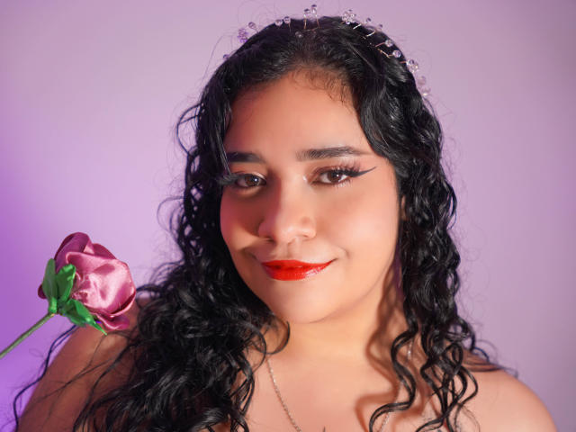 Alicia, con cabello negro rizado, lápiz labial rojo y una rosa rosa en su mano derecha, lleva una diadema brillante, tiene un rostro lindo y atractivo, y una ligera sonrisa..