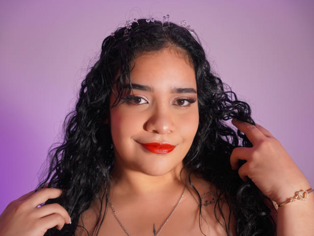 AliciaRelas with curly black hair, red lipstick, and a cute crown, holding her hair, showing off her tits with a silver necklace and a gold bracelet.