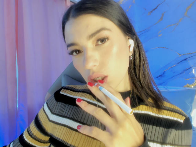 JasmineHypery with red lipstick, holding a cigarette, wearing a striped sweater, and earbuds, has red nails, and is looking seductively at the camera.