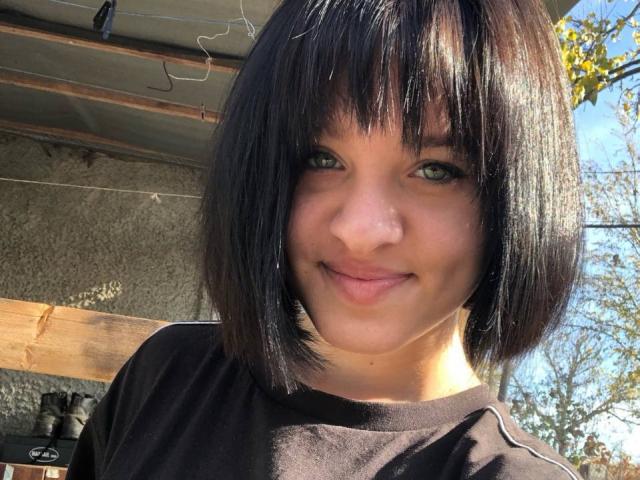 Ssimona with short black hair, green eyes, and a slight smile, wearing a black shirt, sunlight highlighting her face, small nose, and soft skin.