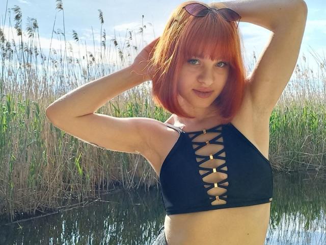 Ssimona with red bob, black lace-up bikini top, sunglasses on head, arms raised, standing by water, showing her tits and armpits.