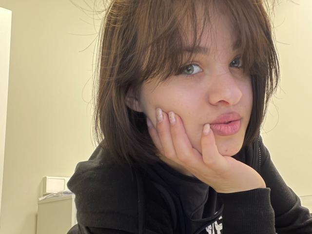 Ssimona with messy brown hair, green eyes, and pink lips, pouting with her hand on her cheek, wearing a black hoodie, showing off her cute nose and small tits.