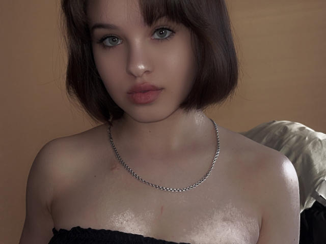 Ssimona has pale skin, short brown hair, and blue eyes. She's wearing a black strapless top and a silver chain necklace. Her tits are slightly visible with small scars.