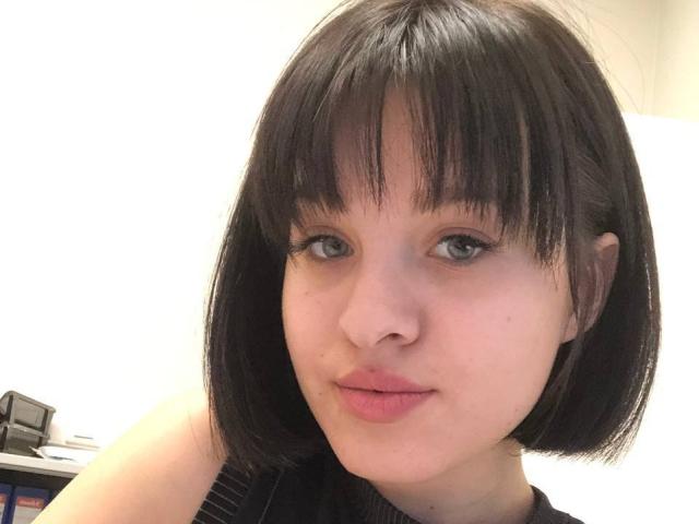 Ssimona has a cute, pale skin, short black bob with straight bangs, blue eyes, and pink lips. She's wearing a black sleeveless top. Her tits are small.