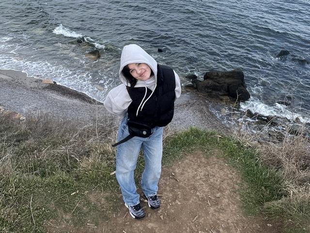Ssimona stands on a grassy path wearing a black vest, white hoodie, blue jeans, and black shoes, with a camera strap, smiling at the camera, by the ocean.
