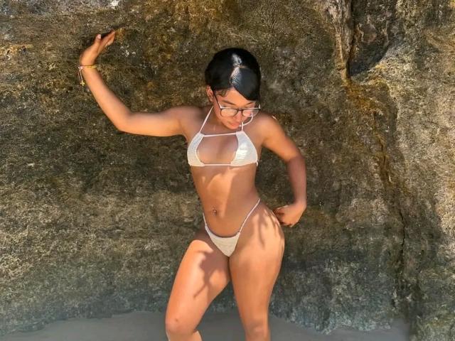 CindiLovely with tan skin, black hair in a ponytail, wearing a white string bikini, standing against a rocky wall, her right arm up, left hand on her hip.