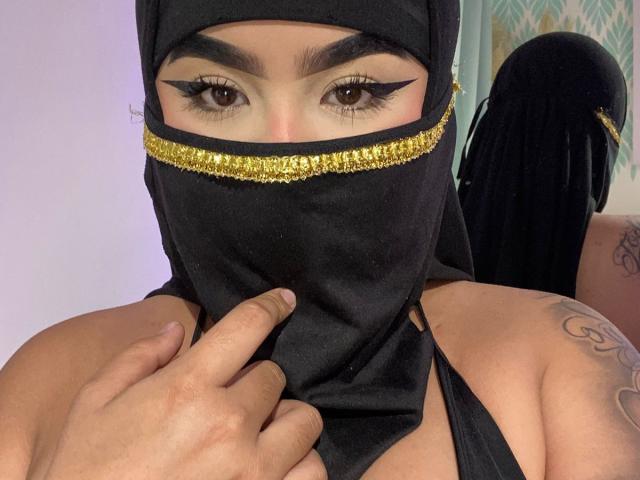 MiaHadid with black mask, gold trim, and thick eyeliner, pointing at her masked mouth, visible tattoo on right shoulder, brown eyes, and tan skin, in a selfie.
