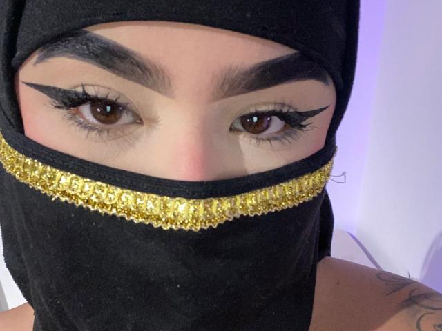 MiaHadid's face with thick black eyebrows, brown eyes, and gold-embellished black mask covering her nose and mouth. She has long, dramatic eyeliner and a tattoo on her right shoulder.