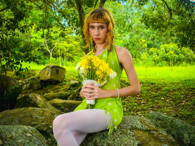AuraFire with blonde hair, green polka-dot dress, white tights, holding yellow and white flowers, sitting on rocks in a green forest, looking serious, hands on bouquet, necklace.