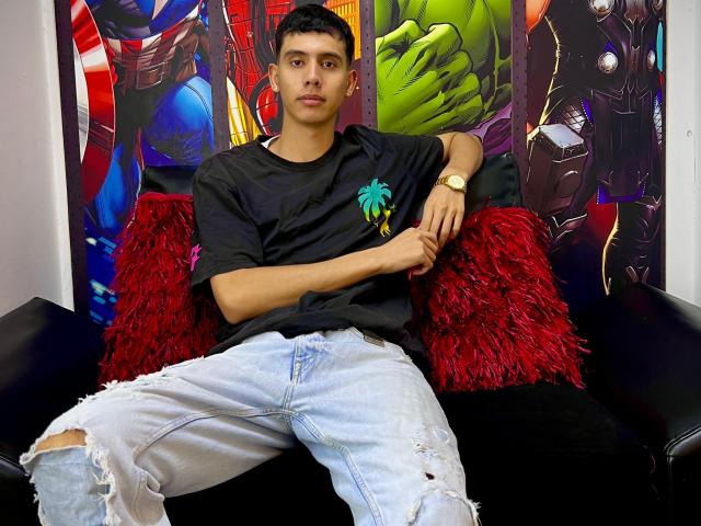 MateoStrogn with short black hair, tan skin, and a serious face, wears a black tee, a palm sticker, ripped jeans, and a gold watch, sitting on a black chair.