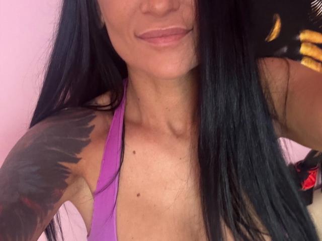 AndraD's face with long black hair, pink lips, and a purple halter top, showing a black tattoo on her right shoulder and a small mole on her chest.