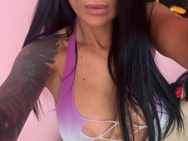AndraD with long black hair, purple gradient sports bra showing her tits with cutout sides, tattooed arm, and pink lips.