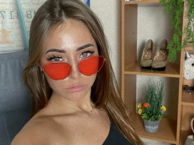 Miliaana, avec des lunettes de soleil rouges, de longs cheveux bruns et un maquillage complet, montrant sa poitrine dans un haut noir, assise dans une pièce avec une étagère en bois et des plantes en pot..