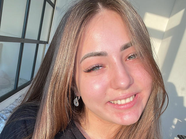 Miliaana has shiny, sunlit skin, long brown hair, green eyes, and a cute smile. She's wearing a black top with a cute diamond earring. Her tits are not visible.