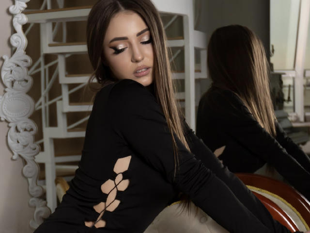 Miliaana with long brown hair, wearing a black long-sleeve top with cut-out designs, leans on a chair, reflected in a mirror, eyes closed, lips slightly parted.