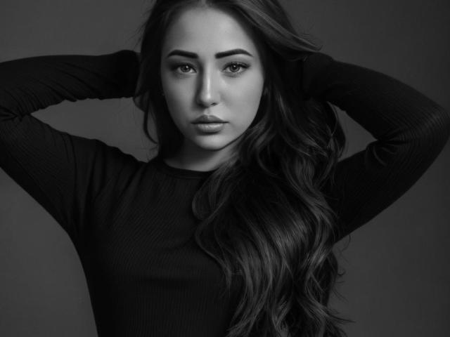 Black-and-white photo of Miliaana with long wavy hair, arms raised, wearing a ribbed black long-sleeve shirt, looking directly at the camera with a serious expression.