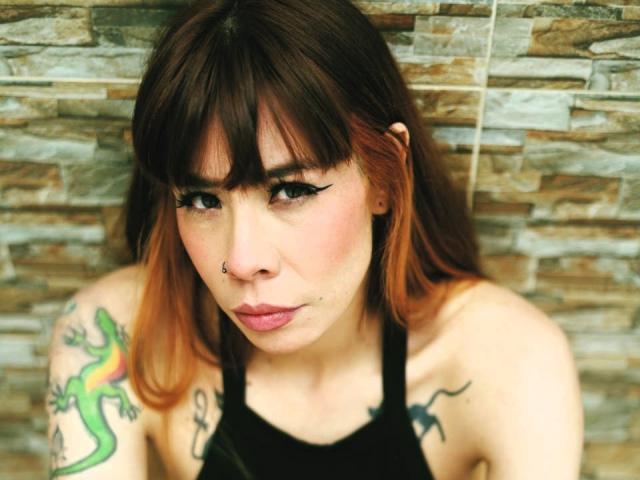 AriiannaHot with brown hair and orange tips, black eyeliner, nose piercing, and tattoos on arms, wearing a black tank top, looking down with a slight pout.