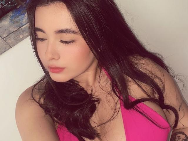 LianaBernocchi has long black hair, pink bikini top, and is looking down with closed eyes. Her tits are slightly visible, and she has a tattoo on her right arm.