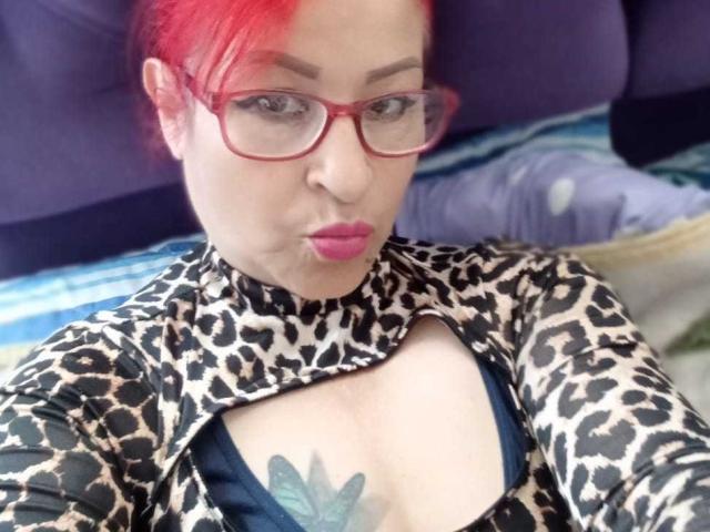 ZafiroMilf with red hair, glasses, pouty pink lips, leopard print top, showing her tits, butterfly tattoo, laying on a bed, looking at camera.