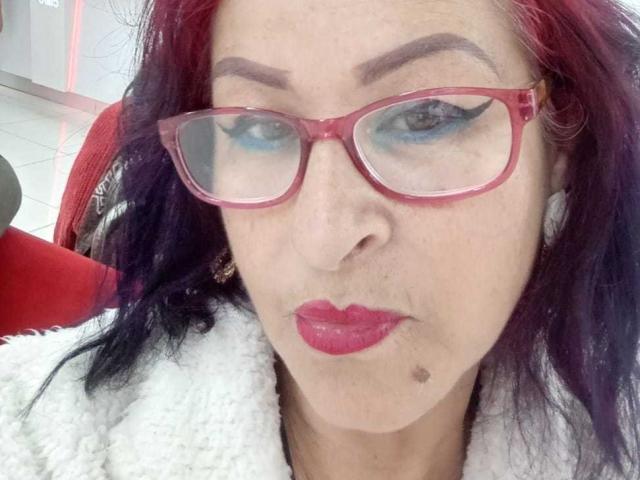 ZafiroMilf with red glasses, blue eyeshadow, purple-black hair, red lipstick, small mole on chin, white fluffy jacket, serious expression.