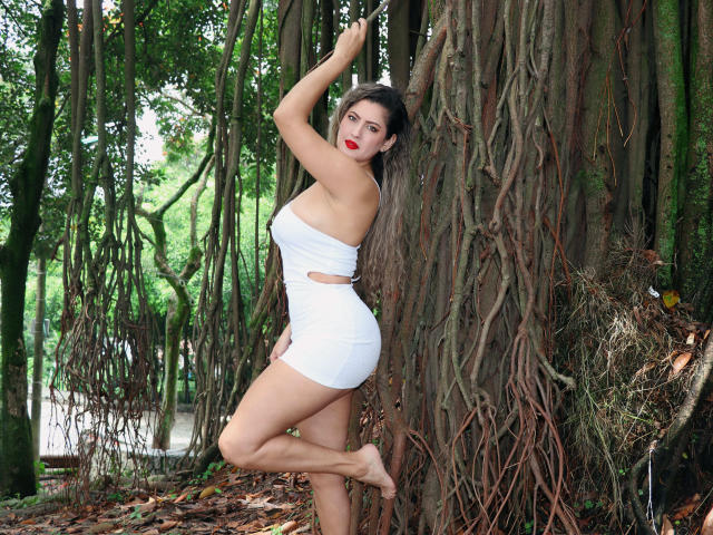 EvaBergman with red lipstick, white strapless cut-out dress, and bare feet, standing against a tree with tangled roots, her right arm raised, showing her curvy ass and tits.